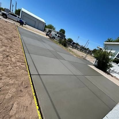 Fresh concrete driveway beside a sandy lot under a clear blue sky, with buildings and parked vehicles nearby