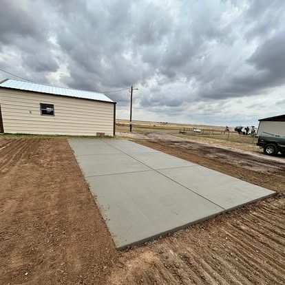 Concrete driveway beside a beige building under a cloudy sky, with a dirt lot and trailer nearby