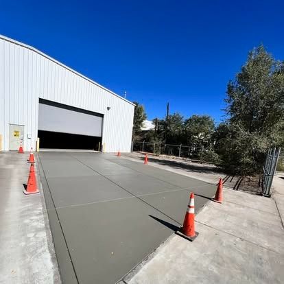 Wide concrete driveway beside white warehouse with open garage door and orange traffic cones under blue sky