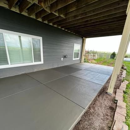 Covered concrete patio beside gray house with white windows and stone edging