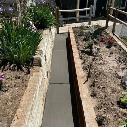 Narrow concrete walkway between a stone wall and a garden bed with young plants.