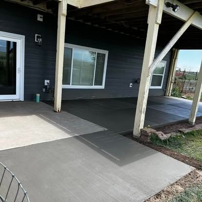 Covered patio with concrete walkway, dark siding, and white support posts beside sliding windows.