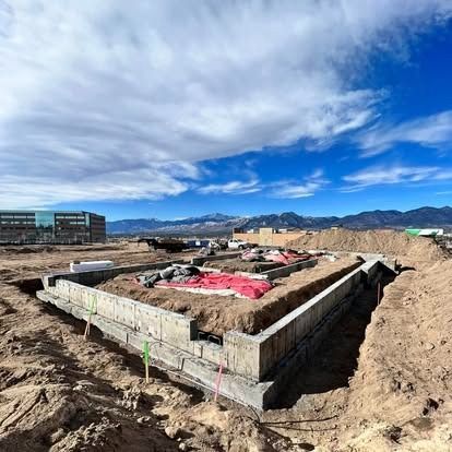 Construction foundation in a dirt lot under a bright blue sky, with buildings and mountains in the background