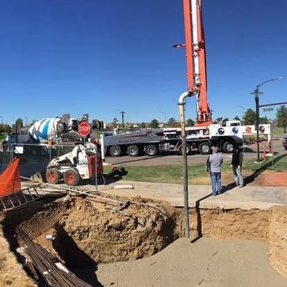 Roadwork site with a concrete pump truck, excavated hole, and workers near a wet concrete pour.