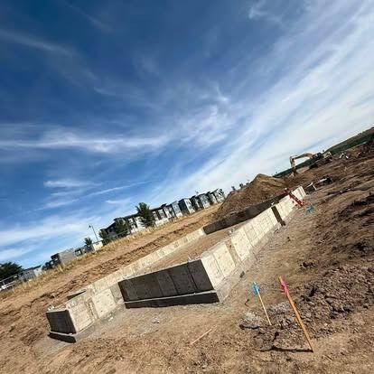 Construction site with concrete foundation walls, dirt piles, and an excavator under a blue sky.