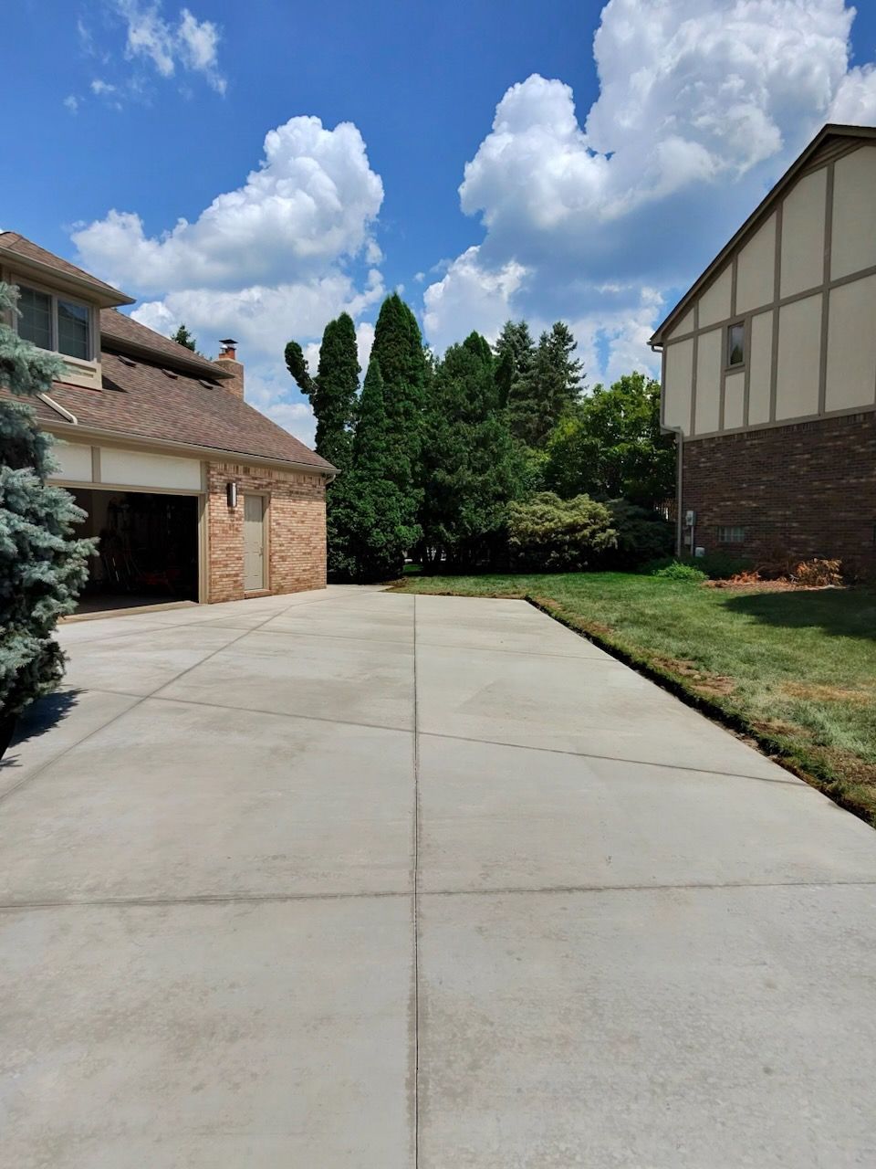 A wide concrete driveway leads between two residential houses toward a lush green tree line under a bright blue sky.