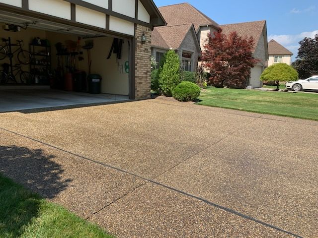 A tan aggregate concrete driveway leads to a suburban house with a two-car garage and a manicured front lawn.