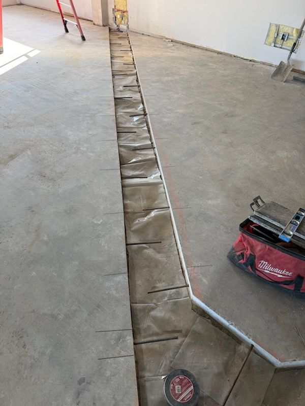 Construction site floor with a long channel cut into the concrete, lined with brown paper and white piping in the center.