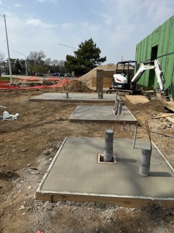 Three square concrete pads with vertical conduits in a construction site with a small excavator near a green building.