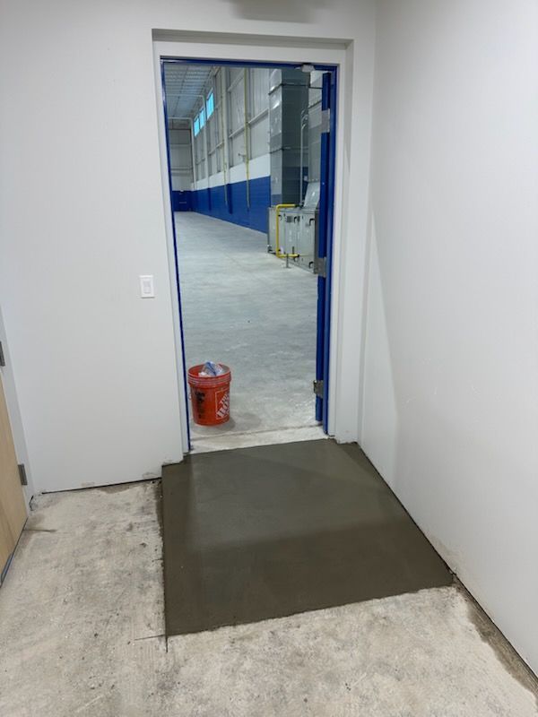 A newly poured concrete ramp leads from a carpeted interior through a doorway into a large, industrial warehouse.