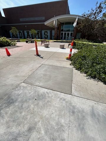 A school entrance with a freshly poured concrete square in the sidewalk, marked by orange safety cones and barriers.
