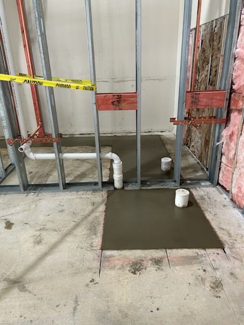 Construction site showing a metal frame wall, plumbing pipes, and fresh concrete patches on a concrete floor.