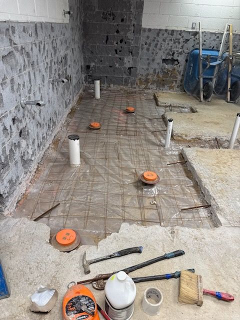 A basement renovation featuring exposed plumbing pipes and a wire mesh grid on plastic sheeting, with tools in the foreground.
