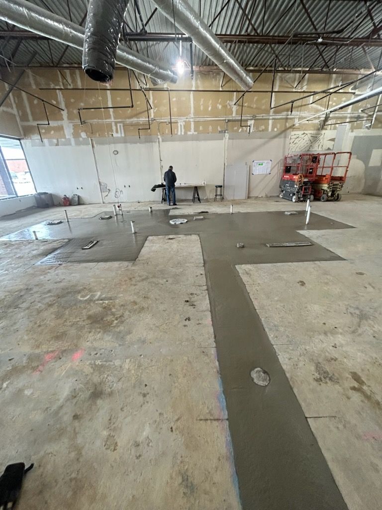 A worker stands in a construction site with newly poured concrete floor patches on an unfinished concrete slab.