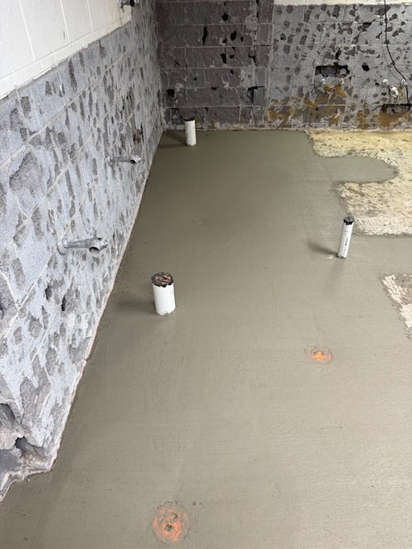 A freshly poured grey concrete floor in a basement with visible white plumbing pipes and rough, unfinished block walls.