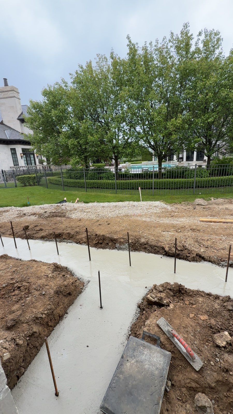A freshly poured concrete foundation trench with vertical rebar stakes, set in a backyard near a house and trees.