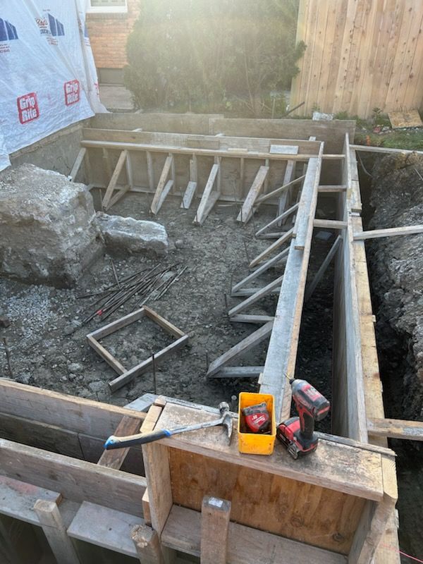 A construction site showing wooden concrete forms for a building foundation set in a dirt trench with tools on the edge.