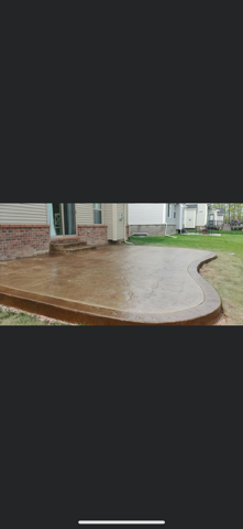 A stamped, curved concrete patio in a backyard adjacent to a house with a brick foundation and a glass sliding door.