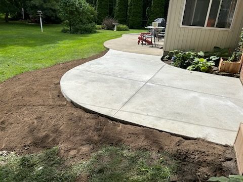 A curved concrete patio edge next to a patch of bare, excavated dirt in a backyard with green grass and trees.