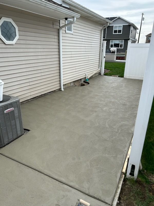 Newly poured concrete patio next to a beige-sided house with an HVAC unit and a white vinyl fence in the background.