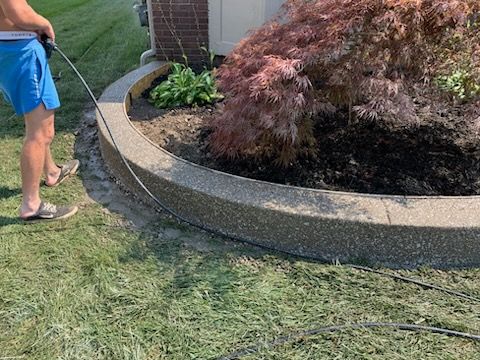 A person in blue shorts uses a pressure washer to clean a curved concrete retaining wall near a garden bed and lawn.