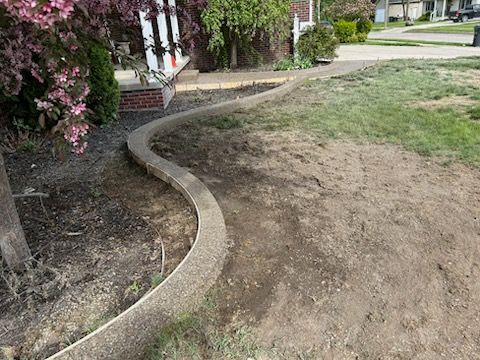 A curved concrete landscape border separates a mulched garden bed from a patch of bare soil in a front yard.