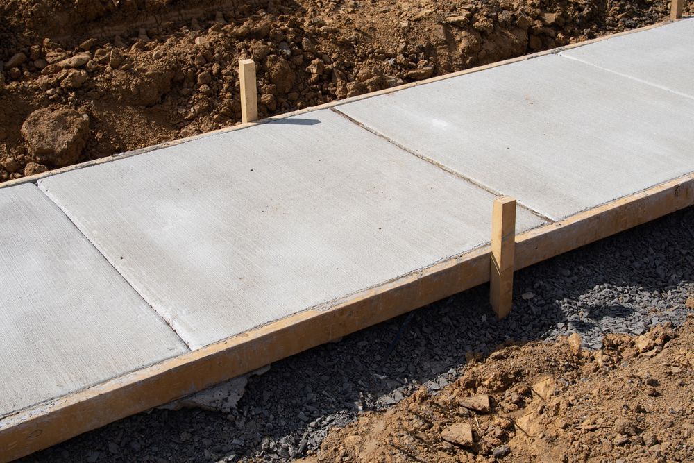 A newly poured concrete sidewalk with wooden form boards held in place by stakes against a dirt embankment.