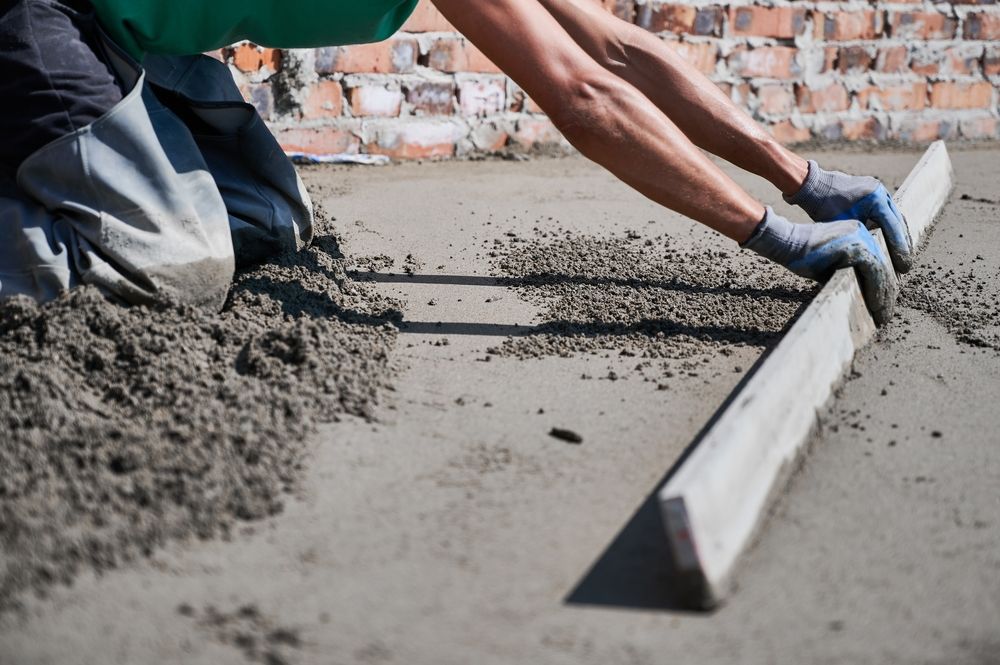 A worker kneeling on a floor uses a long wooden leveling tool to smooth freshly laid concrete.