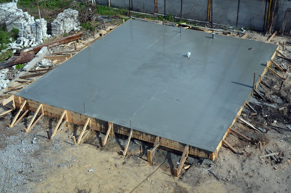 An aerial view of a freshly poured square concrete foundation slab with wooden formwork on a construction site.