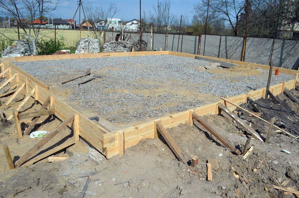 Wooden formwork surrounds a rectangular foundation filled with gravel for a construction project on a sunny day.