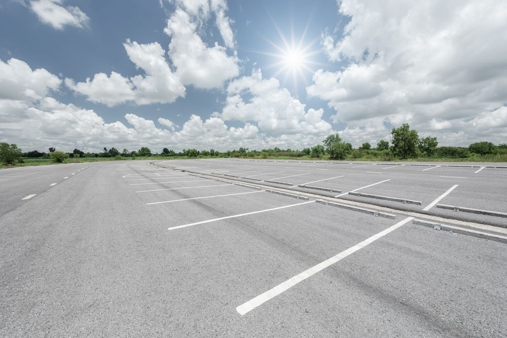 Empty outdoor parking lot with painted white lines under a bright blue sky with scattered clouds and visible sun.