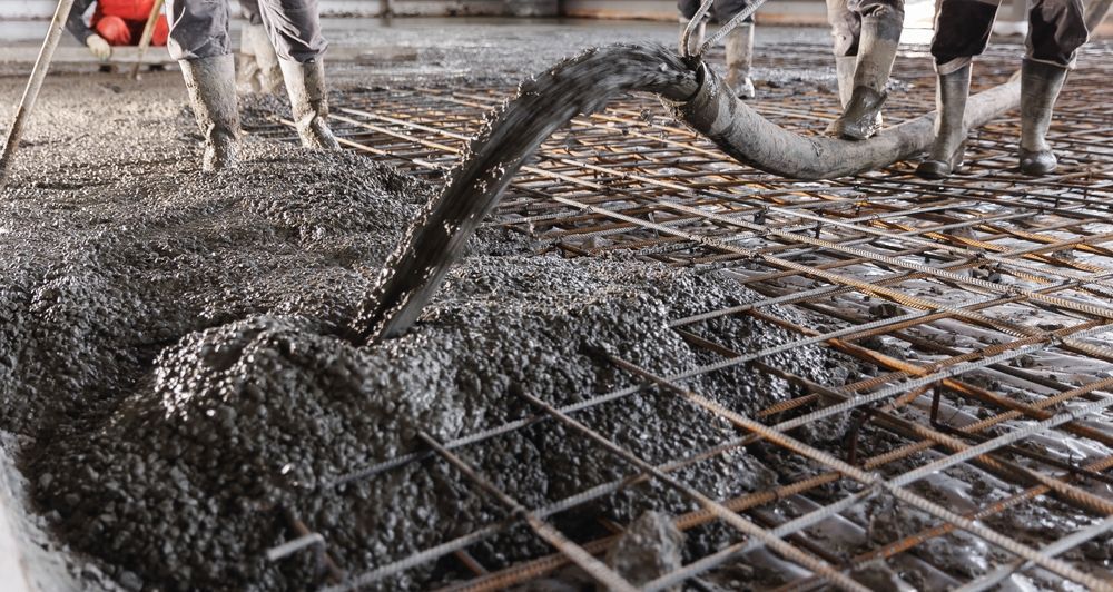 Workers pour wet concrete from a hose over a wire mesh grid on a construction site.