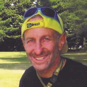 Man smiling, wearing a Brazil cap and sunglasses on his head, against a park background.