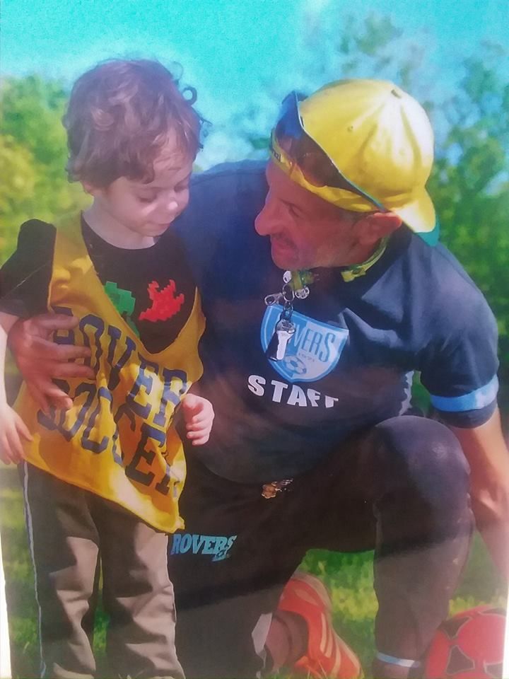 Man in yellow hat kneels, arm around child wearing a yellow vest. Both look down, outdoors.