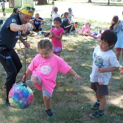Children play soccer with an instructor in a park. One kicks a ball. Others watch, some in pink shirts.