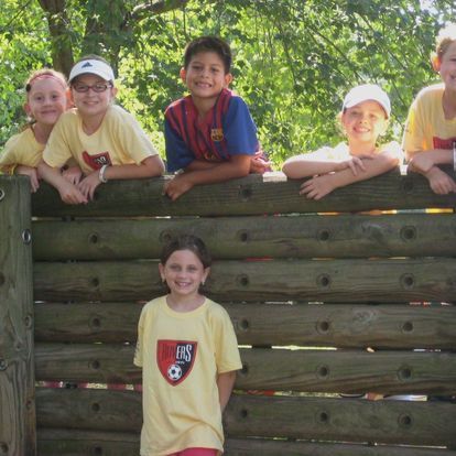 Children in yellow shirts and hats smiling, leaning on a wooden structure; one child stands in front.