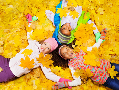 Three smiling children lying in yellow autumn leaves, holding up colorful leaves and looking at the camera