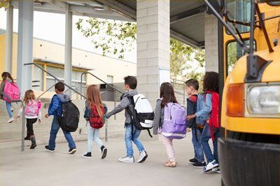 Children with backpacks walking under a covered school entrance beside a yellow bus