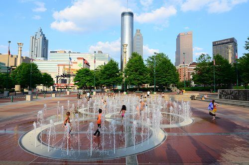 City fountain plaza with children playing in water, trees, and tall buildings in the background.
