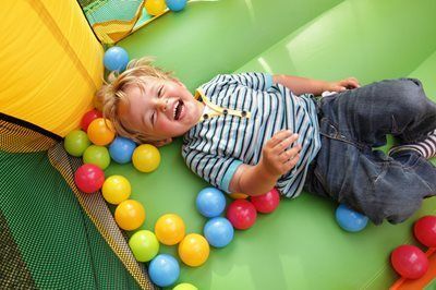 Child laughing on a green play mat among colorful balls near a yellow slide