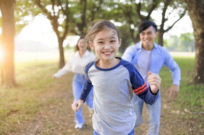 Smiling child running toward the camera in a park, with two adults following behind among trees.