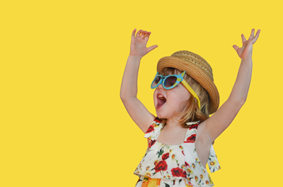 Child in sunglasses and straw hat with arms raised against a bright yellow background