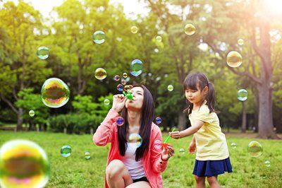 Two children blowing bubbles in a sunny park, with green trees in the background