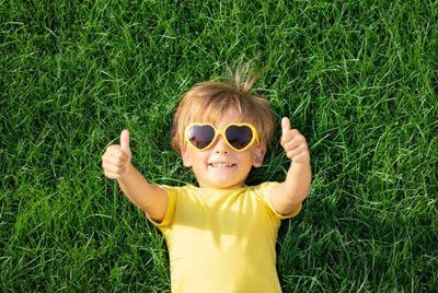 Child in yellow shirt and heart-shaped sunglasses lying on green grass with both thumbs up