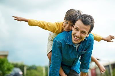 Smiling man carrying a child on his back in a green field, both with arms outstretched