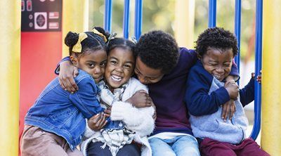 Four children in blue jackets cuddle and smile on a playground bench