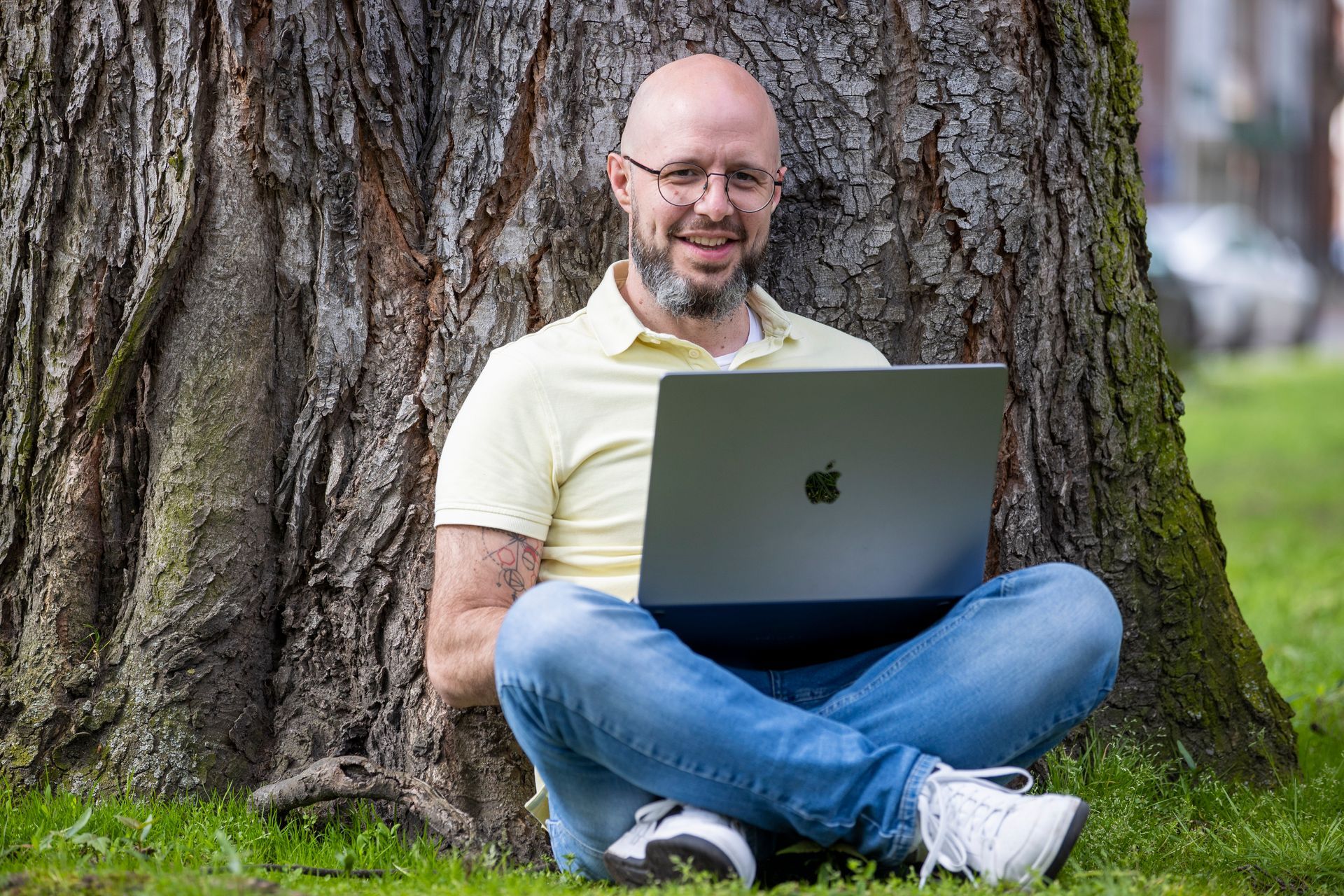 Een man zit lachend in het gras tegen een boom terwijl hij buiten een laptop gebruikt.