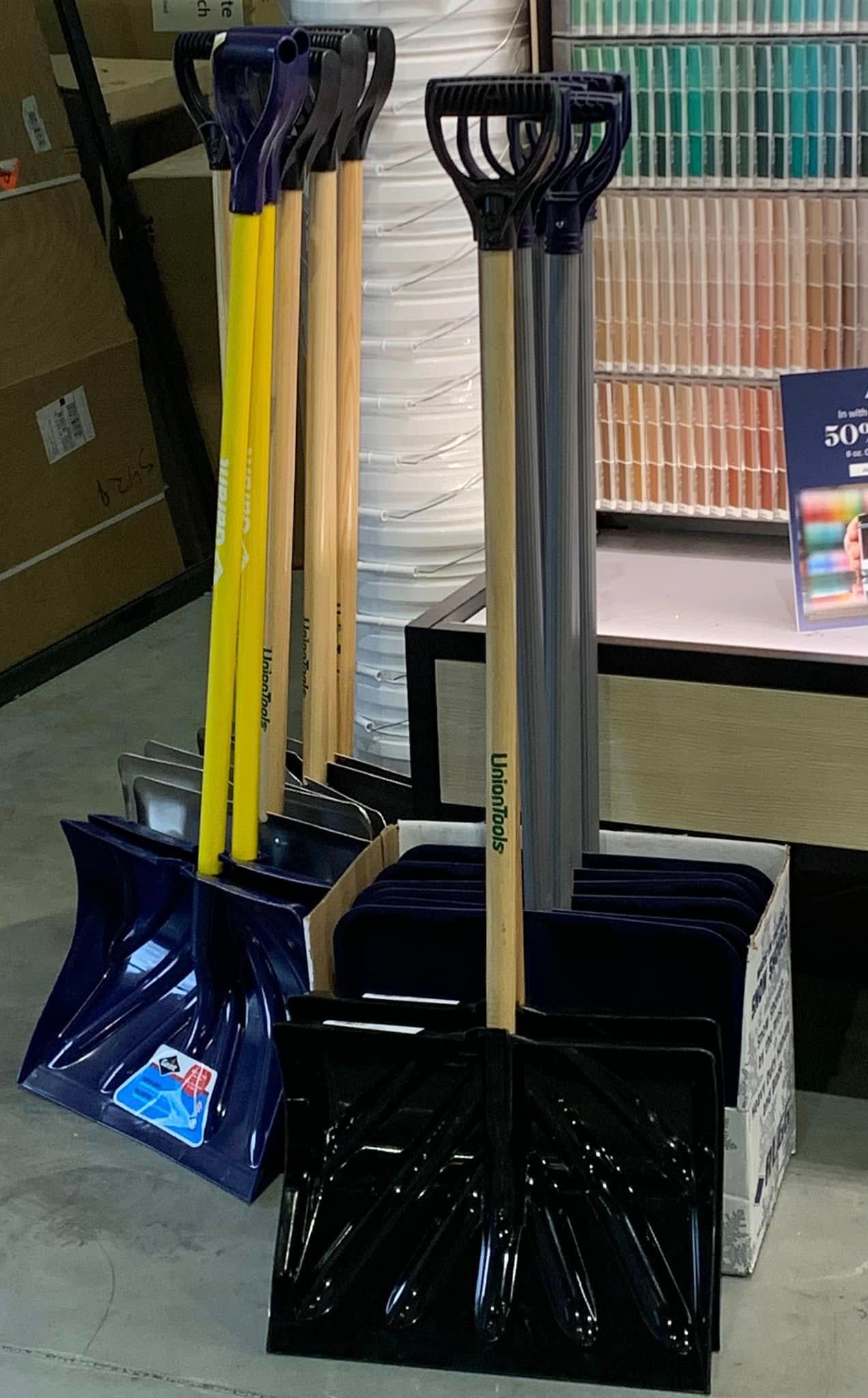 Snow shovels and a black snow pusher displayed upright indoors near a window.