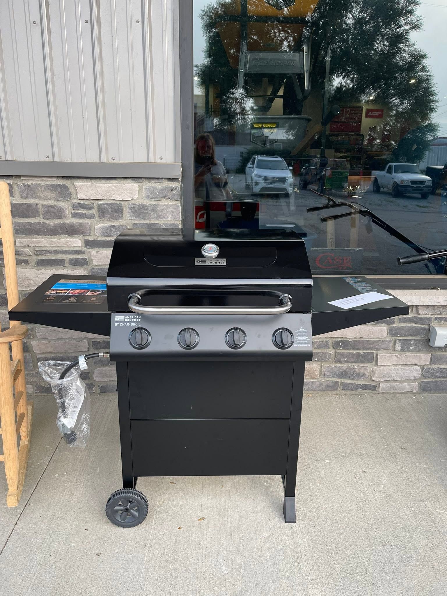 Black three-burner gas grill on a sidewalk outside a storefront, with side shelves open.