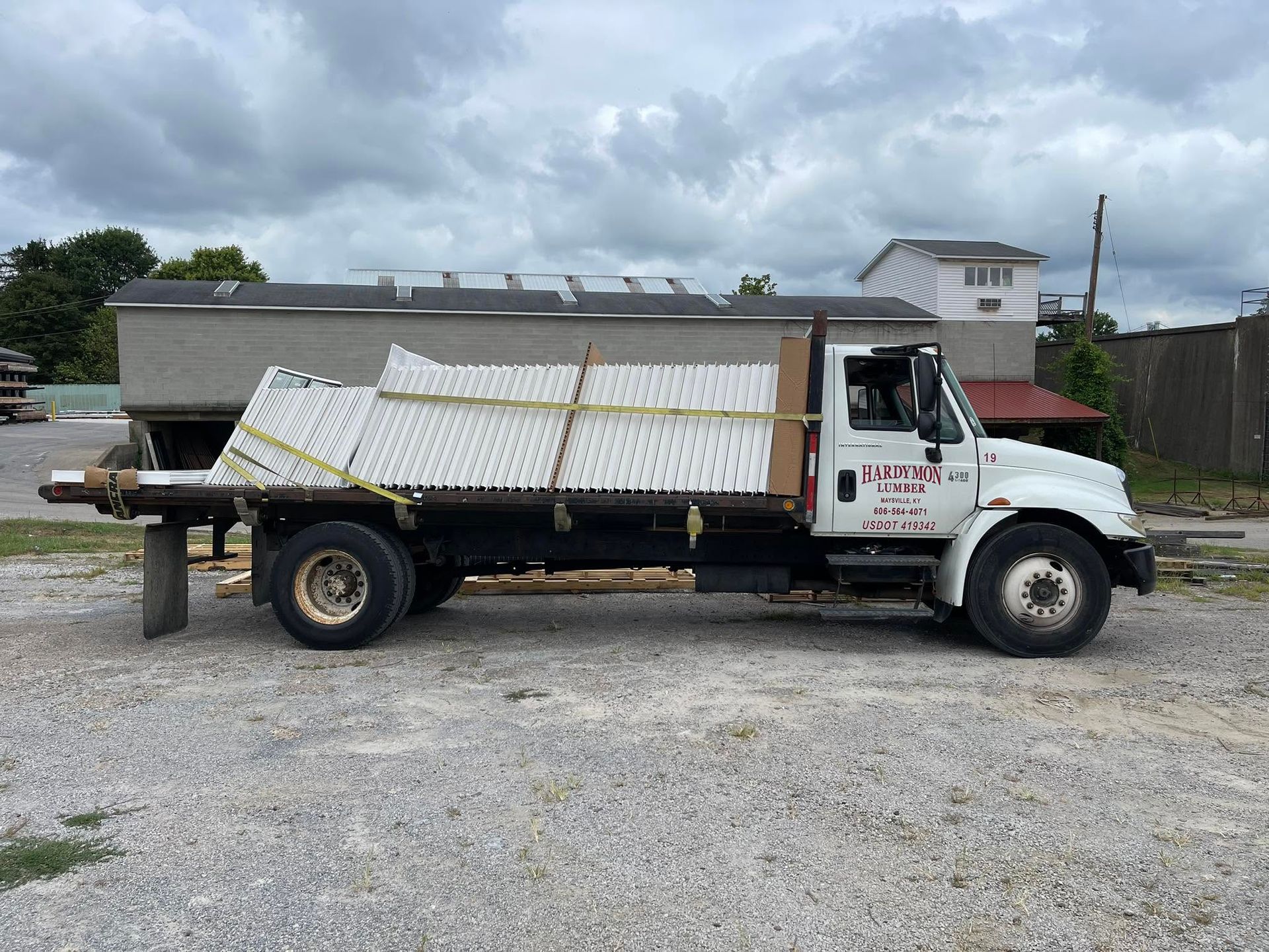 White flatbed truck loaded with stacked white panels, parked on gravel under cloudy skies.